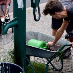 Ein Foto von einem öffentlichen Brunnen und einem Mann, der mit einer Gießkanne Wasser aus einer Schubkarre schöpft.