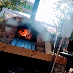 Ein Foto om fertigen Steinbackofen im Garten. Die Klappe ist offen, drinnen lodert das Feuer. Neben dem Ofen steht der Einschieber aus Holz.