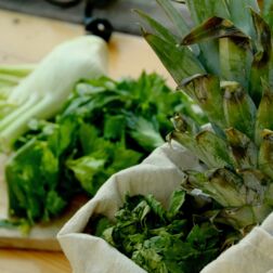 Ein Foto von verschiedenem Obst und Gemüse: Ananas, Fenchel und Kräuter. Ein Teil ist in einem Stoffbeutel, ein Teil liegt auf einem Holzbrett auf einem Tisch.