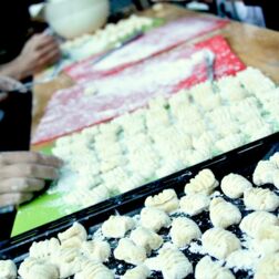 Ein Foto von einem langen Tisch auf dem ein Blech und zwei Schneidebretter mit fertig geformten, rohen Gnocci stehen. Außerdem Hände, die die Gnocci formen.