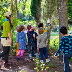 Eine Kindergruppe_hängt Infos zum Thema Stadtnatur zwischen zwei Bäumen auf. Sie werden von einer Naturpädagoin begleitet.