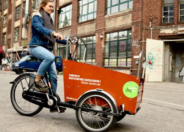 Eine Frau fährt ein Lastenfahrrad der flotte-berlin.de im Hintergrund ein Fabrikhaus