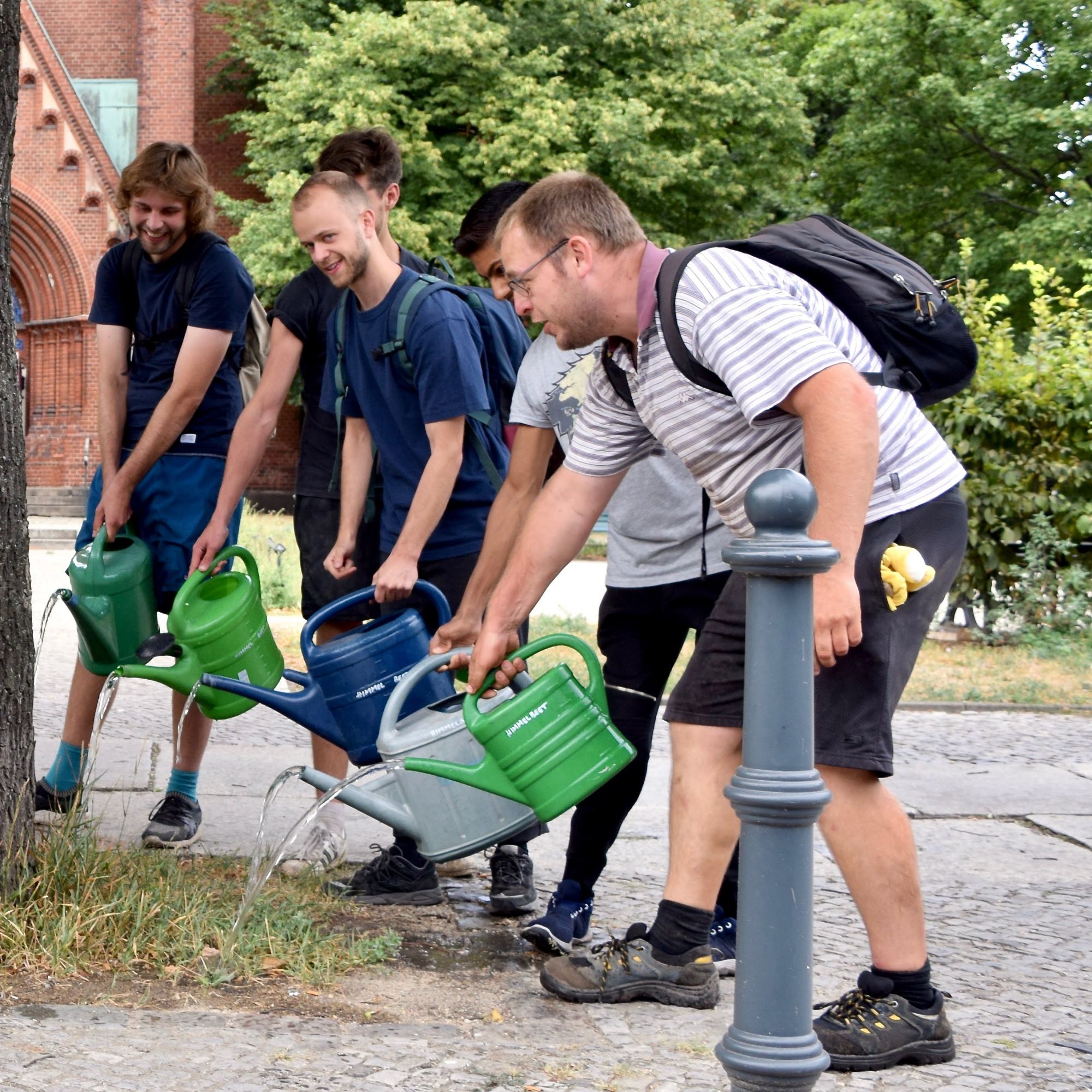 Ein Foto von einer Gruppe Menschen im Halbkreis mit Gießkannen in den Händen Gemeinsam wässern sie einen Stadtbaum.